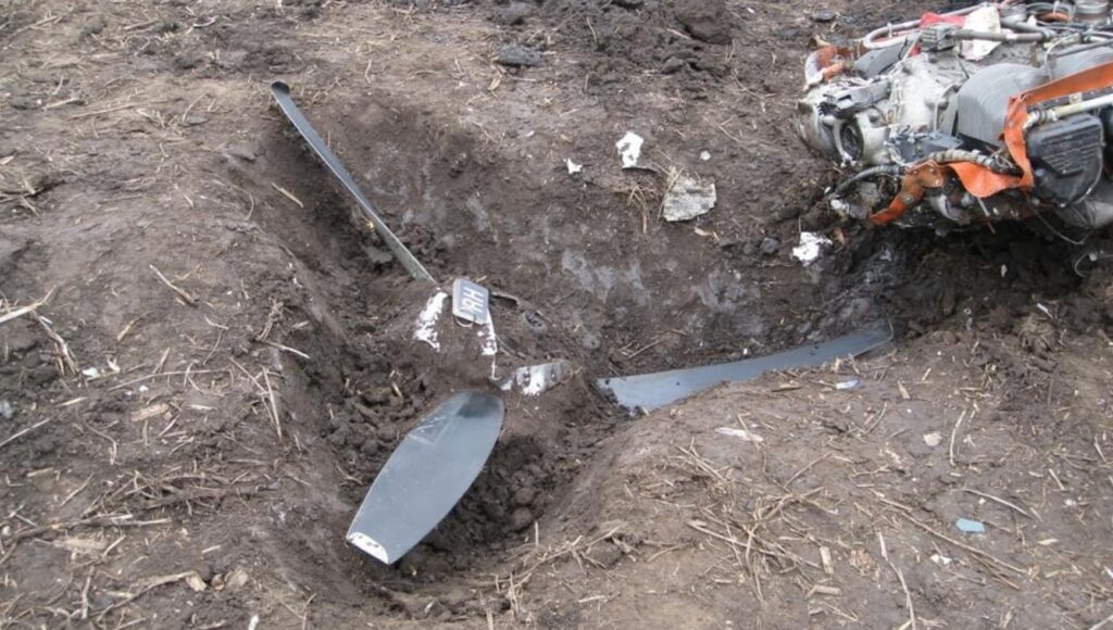 Close-up of a Cessna 414A propeller blade and engine partially buried in a crater, showing impact damage to the engine assembly and surrounding soil.
